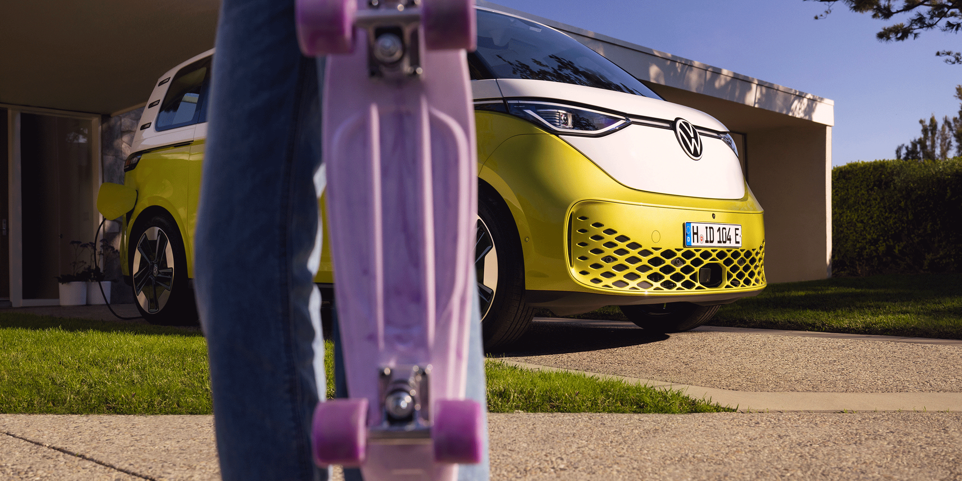 O ID. Buzz estacionado em frente de um acesso; em primeiro plano uma rapariga com uma prancha de skate roxa. 
