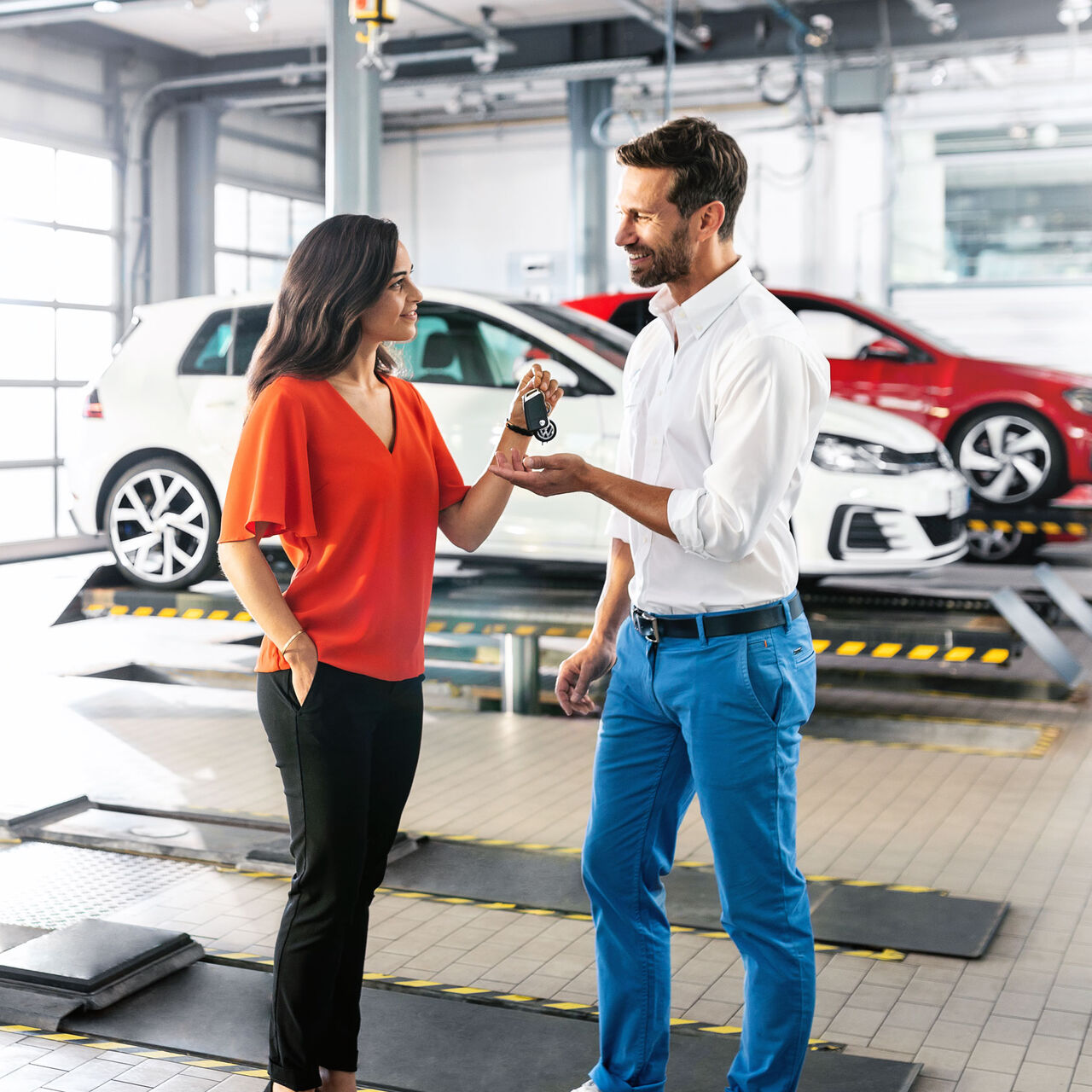 Eine Frau übergibt Ihren Autoschlüssel in einer Autowerkstatt an einen Mann. Im Hintergrund sind Volkswagen zu sehen.
