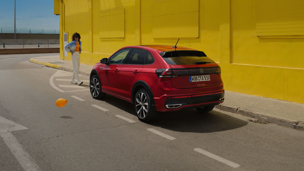 VW Taigo em vermelho na berma da estrada, em frente a um edifício amarelo, vista traseira e lateral, uma mulher dirige-se para o veículo