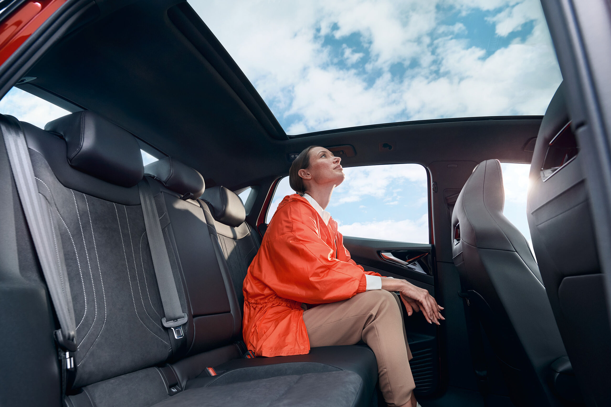 Uma mulher jovem sentada no banco traseiro de um VW ID.5 GTX vermelho a olhar pelo teto panorâmico para o céu azul.