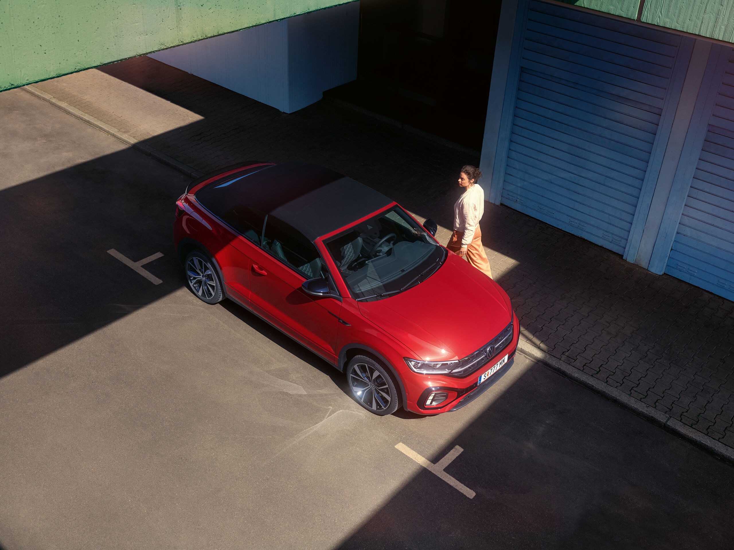 Vista aérea do VW T-Roc Cabrio vermelho com o tejadilho aberto. Estacionado à beira da estrada, uma mulher passa ao lado. 