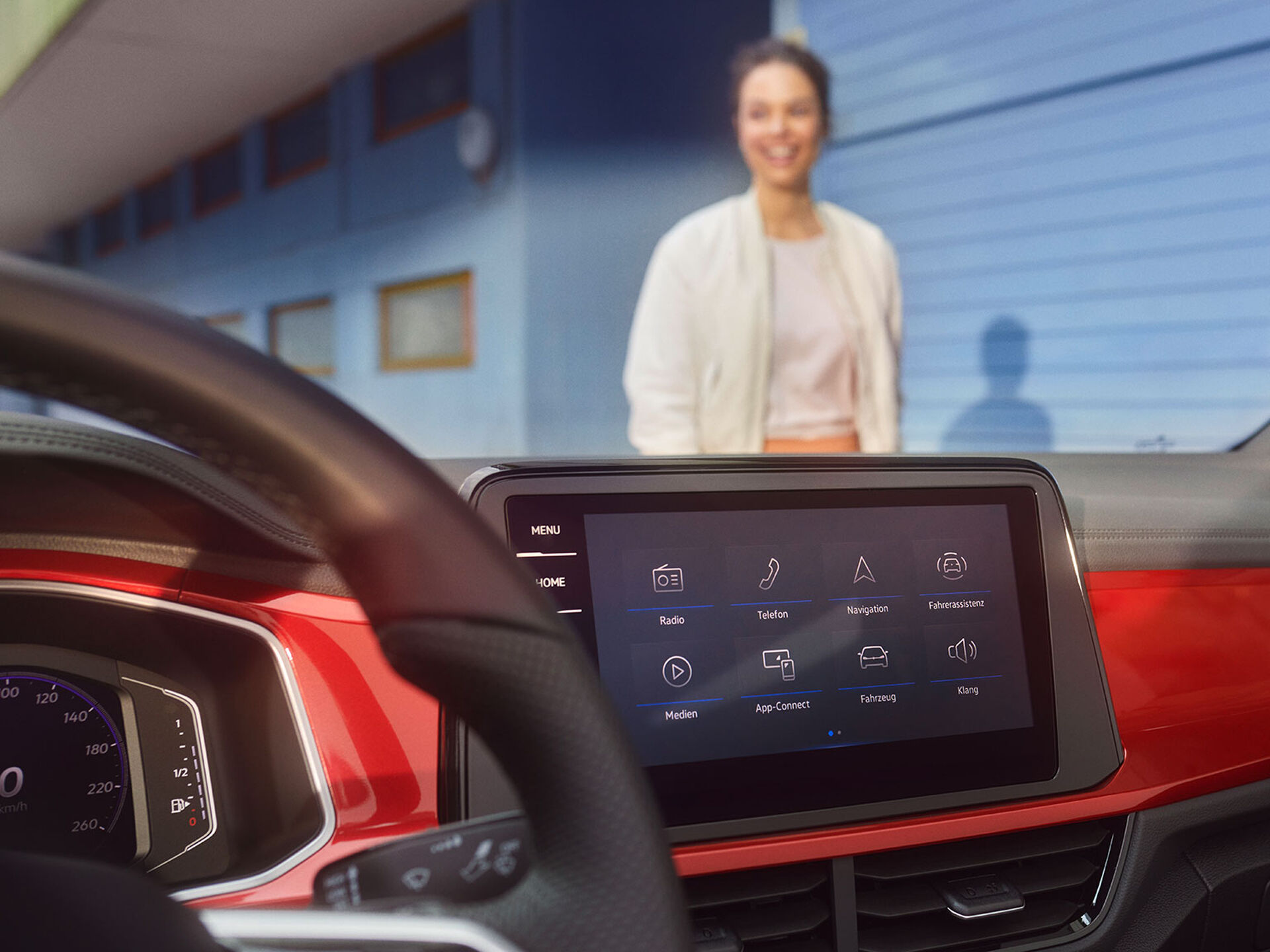 Interior do T-Roc Cabrio: Vista do ecrã com a exibição do menu e o volante multifunções. Uma mulher está de pé, à frente do Cabrio.