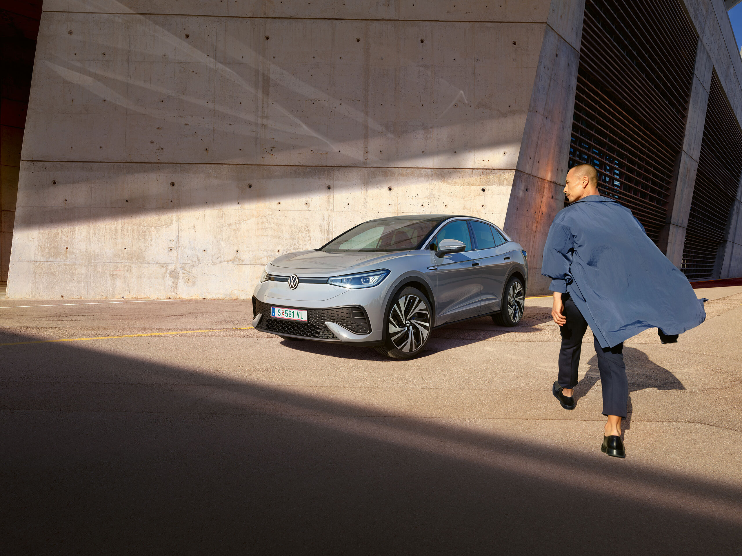 VW ID.5 cinzento estacionado em frente da parede clara de uma casa, vista de frente oblíqua para a direita - um homem dirige-se a passo célere para o automóvel elétrico.