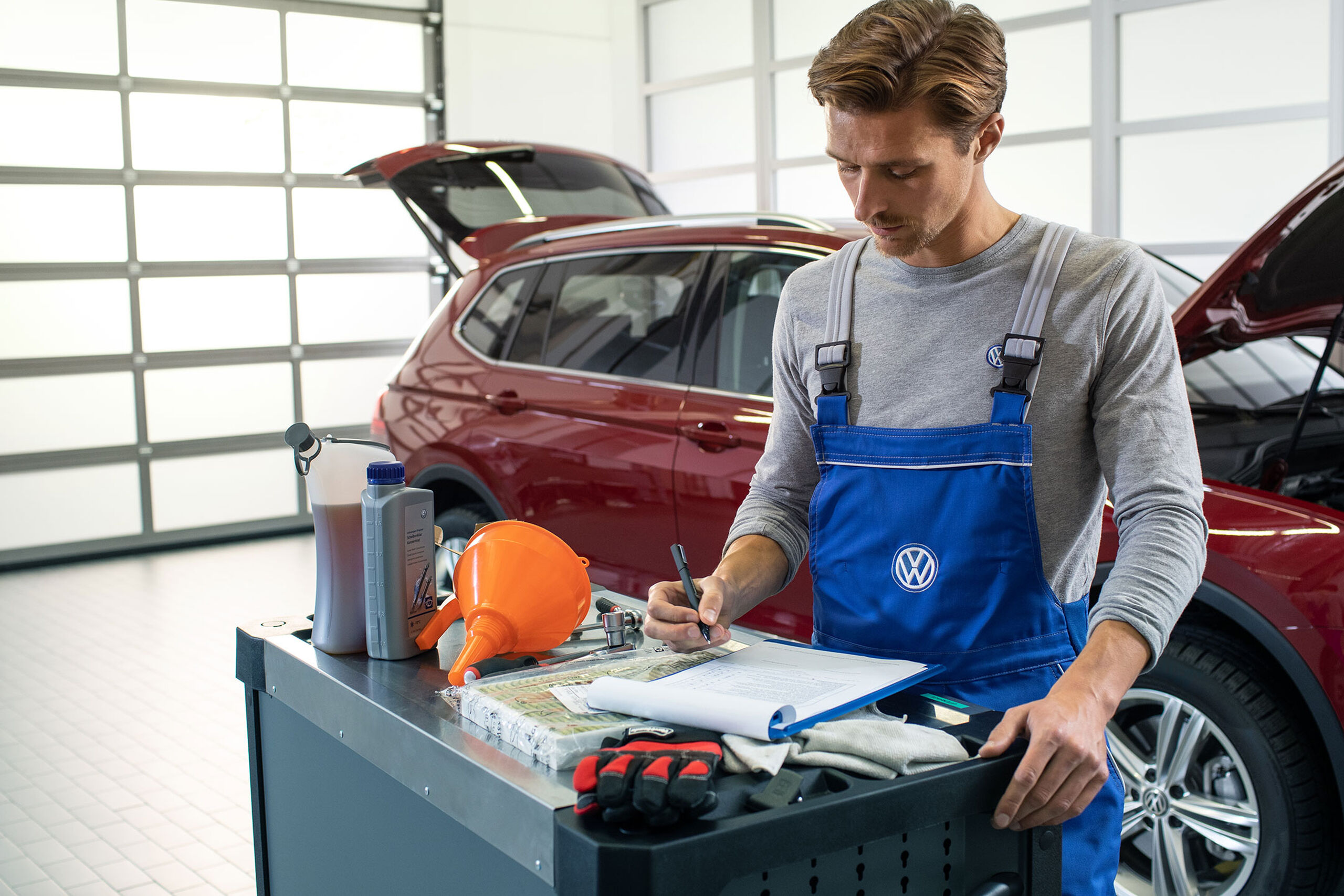 Um empregado do serviço VW está a consultar uma lista de verificação em frente de um VW Tiguan Allspace vermelho com capota aberta e compartimento de bagagem