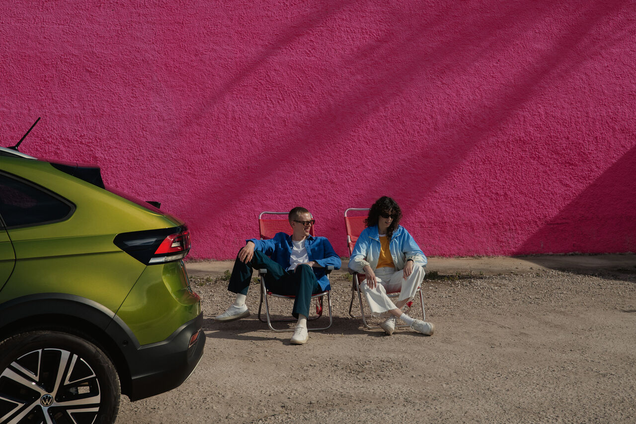 Traseira de VW Taigo verde; atrás duas pessoas sentadas em cadeiras de campismo
