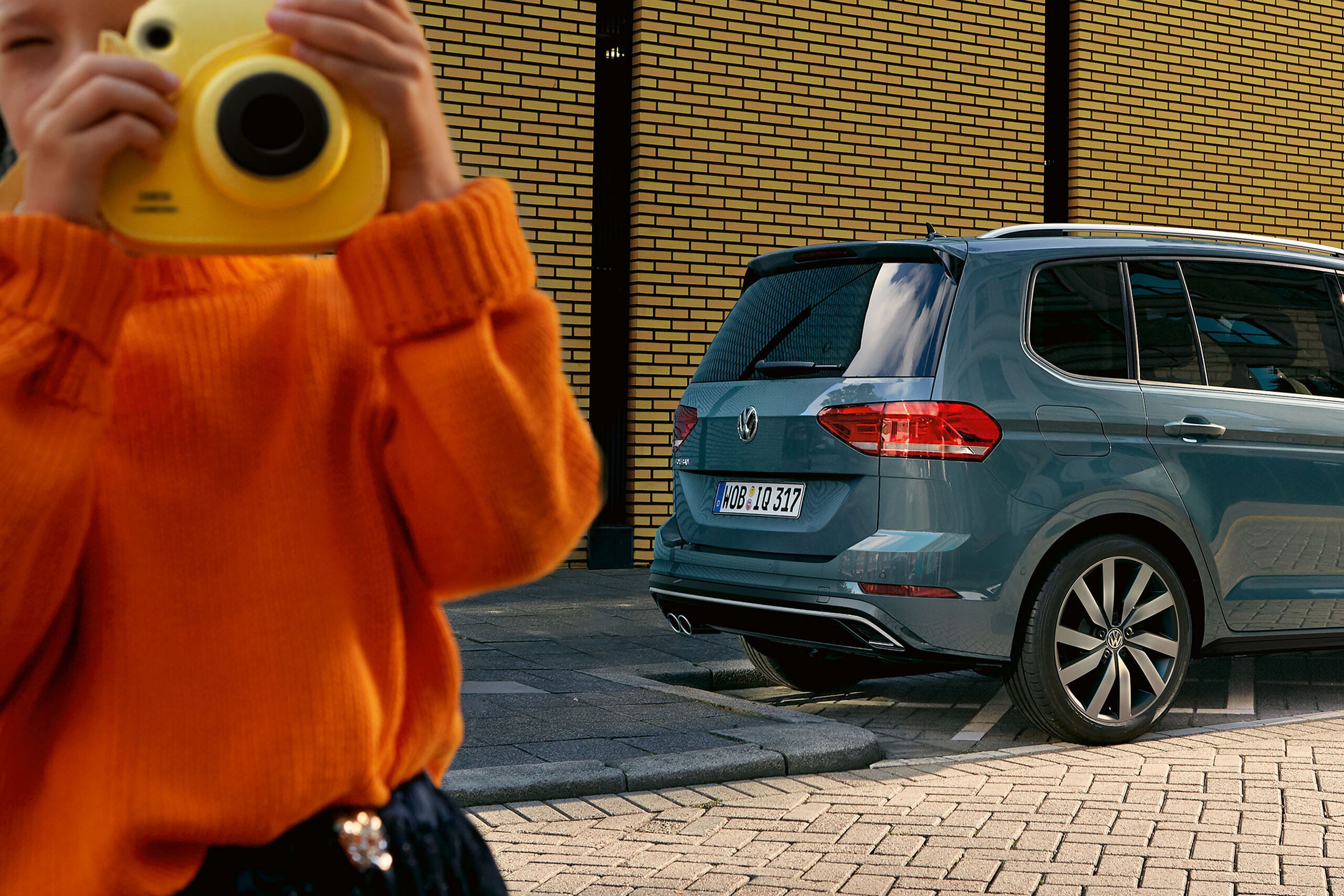 Uma criança com uma grande máquina fotográfica amarela fica em frente de um carro VW azul com pneus de Verão