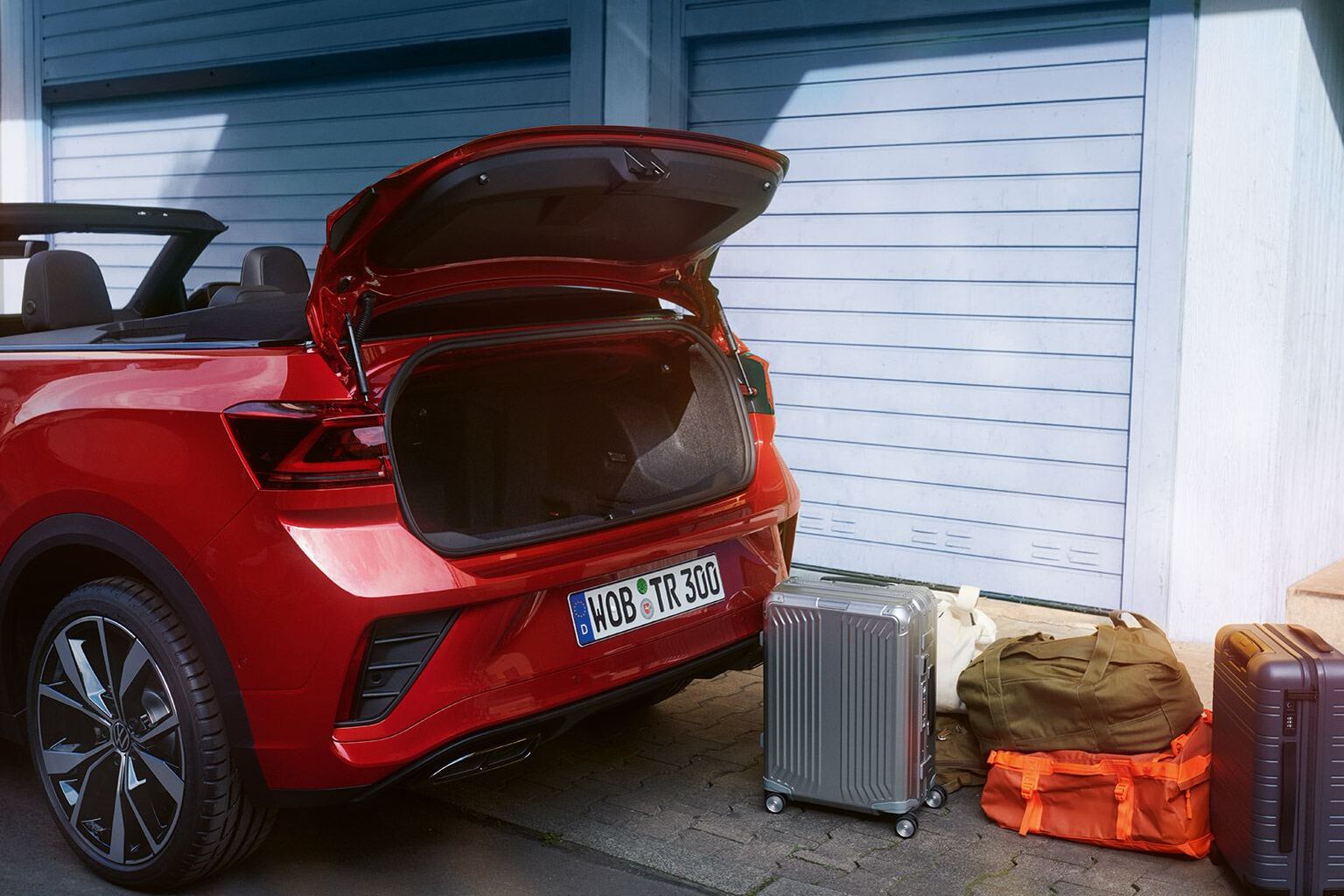 Ein VW T-Roc Cabriolet in rot parkt am Straßenrand. Blick seitlich auf den geöffneten Kofferraum, es stehen zwei Koffer und zwei Reisetaschen daneben. 