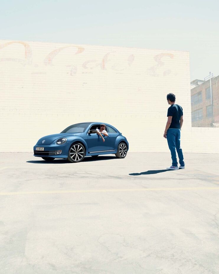 Blauer VW Beetle in der Seitenansicht vor einer Mauer. Daneben steht ein Mann.