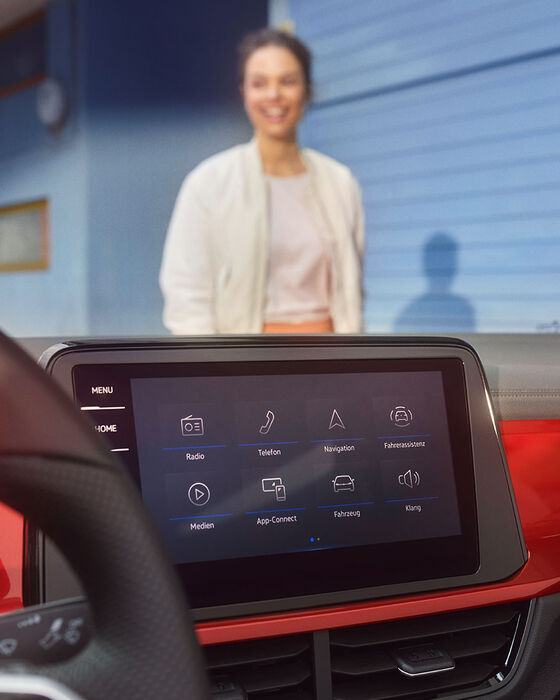 Interior do T-Roc Cabrio: Vista do ecrã com a exibição do menu e o volante multifunções. Uma mulher está de pé, à frente do Cabrio.