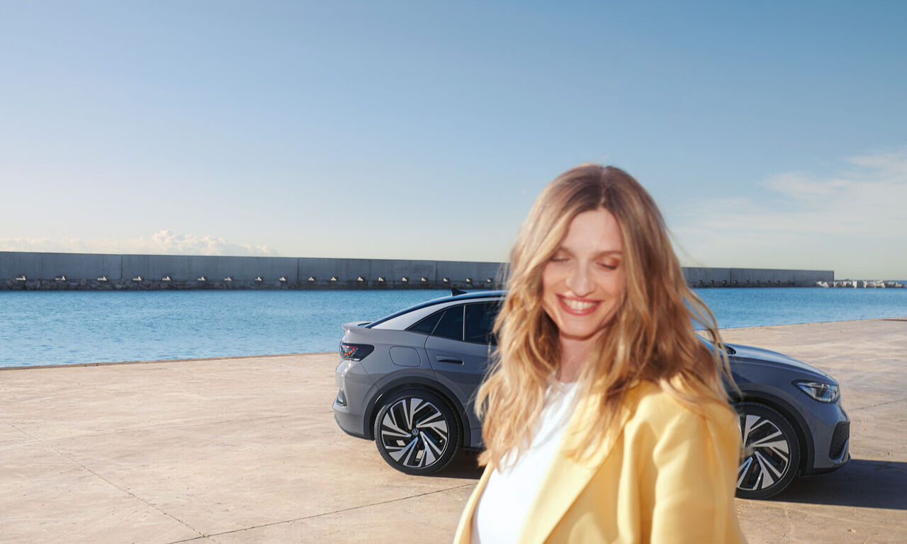 Mulher jovem a sorrir em frente de um VW ID.5 cinzento estacionado num porto.