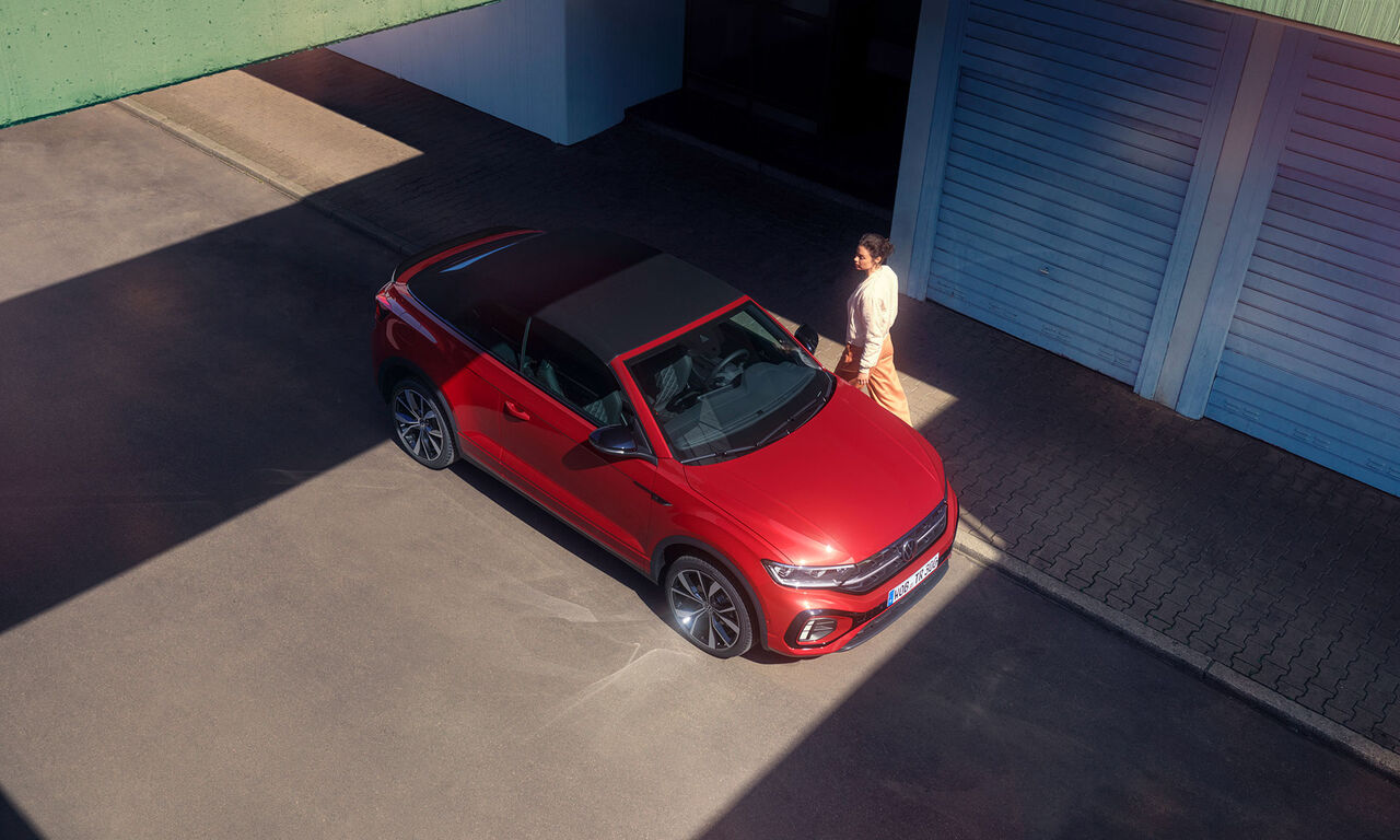 Vogelperspektive auf das rote VW T-Roc Cabriolet mit geöffnetem Dach. Parkend am Starßenrand, eine Frau geht vorbei. 