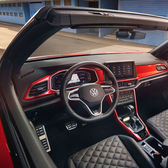 Interior do VW T-Roc Cabrio: Vista de cima para o cockpit, através do tejadilho aberto, com elementos decorativos em vermelho e os bancos da frente. 