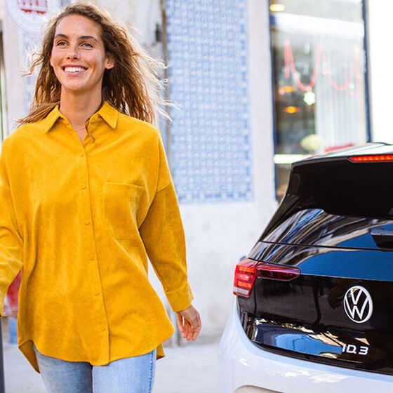 Eine Frau in einer gelben Bluse geht lächelnd von ihrem VW ID.3 weg