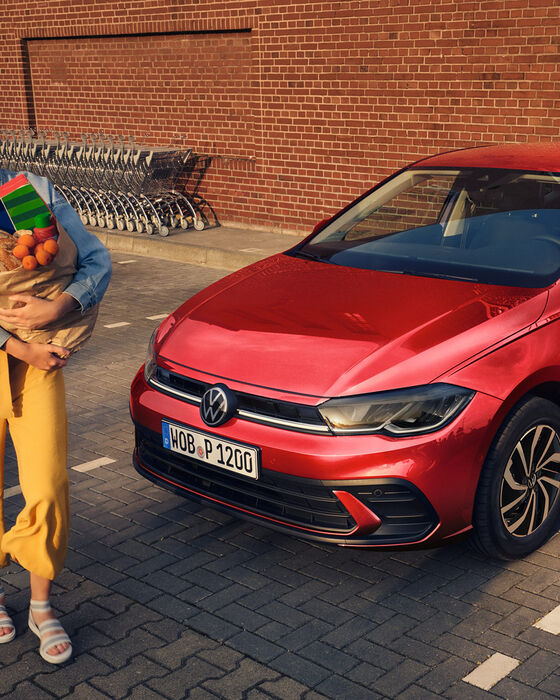 Polo Life vermelho num parque de estacionamento, vista oblíqua da dianteira. Uma mulher passa a rir, com um saco de compras no braço.