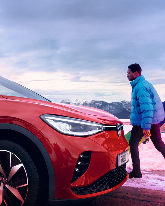 VW ID.4 GTX em vermelho de lado numa estrada na neve. Homem com casaco de Inverno e prancha de snowboard na mão caminha em direção ao SUV desportivo 100% elétrico. 