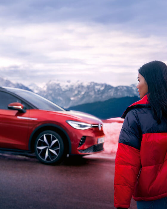 VW ID.4 GTX em vermelho na estrada numa paisagem de montanhas com neve. Mulher de casaco vermelho e preto atravessa a estrada com esquis e olha para a esquerda.