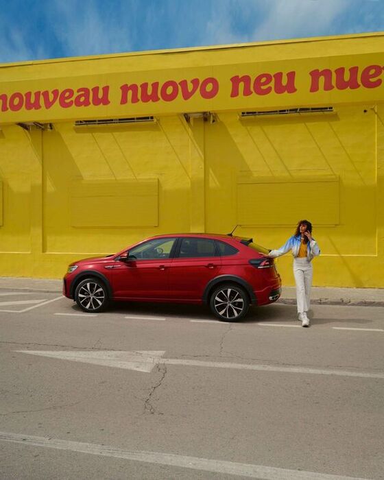 Vista lateral de um VW Taigo vermelho, com o pacote de design opcional “Black Style”, parado junto ao passeio da rua, frente a um edifício amarelo; mulher, de pé, junto do automóvel