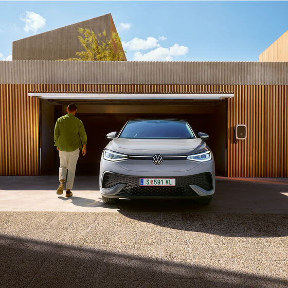 Vista frontal de um VW ID.5 prateado em frente de uma garagem aberta com wallbox, um homem dirige-se para o portão da garagem.