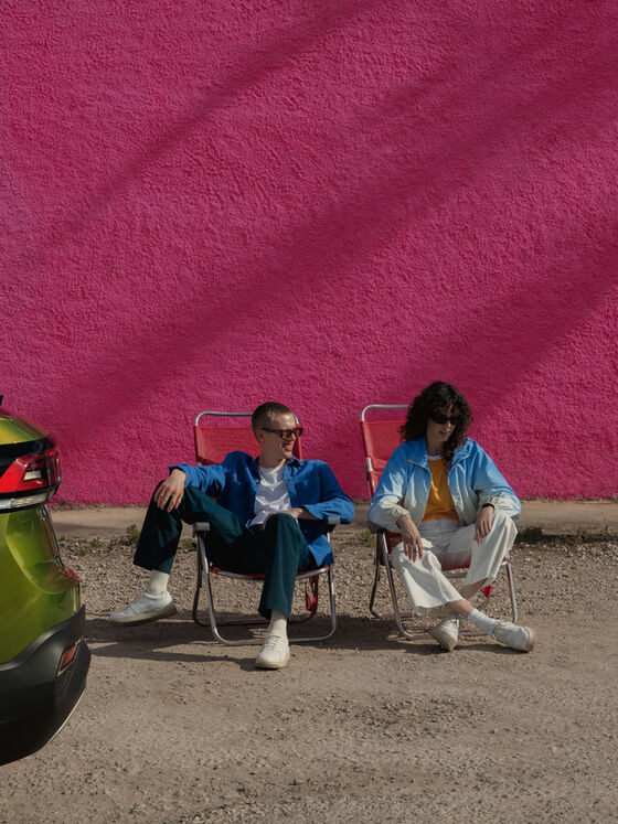 Traseira de VW Taigo verde; atrás duas pessoas sentadas em cadeiras de campismo